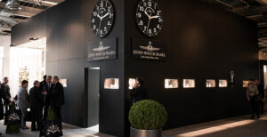 Zeno-Watch Basel trade show booth with two large wall clocks, black walls, and groups of attendees chatting.