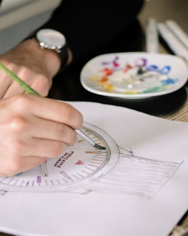 Hands sketching a curved architectural drawing with a protractor and pencil on paper; a paint palette sits in the background.